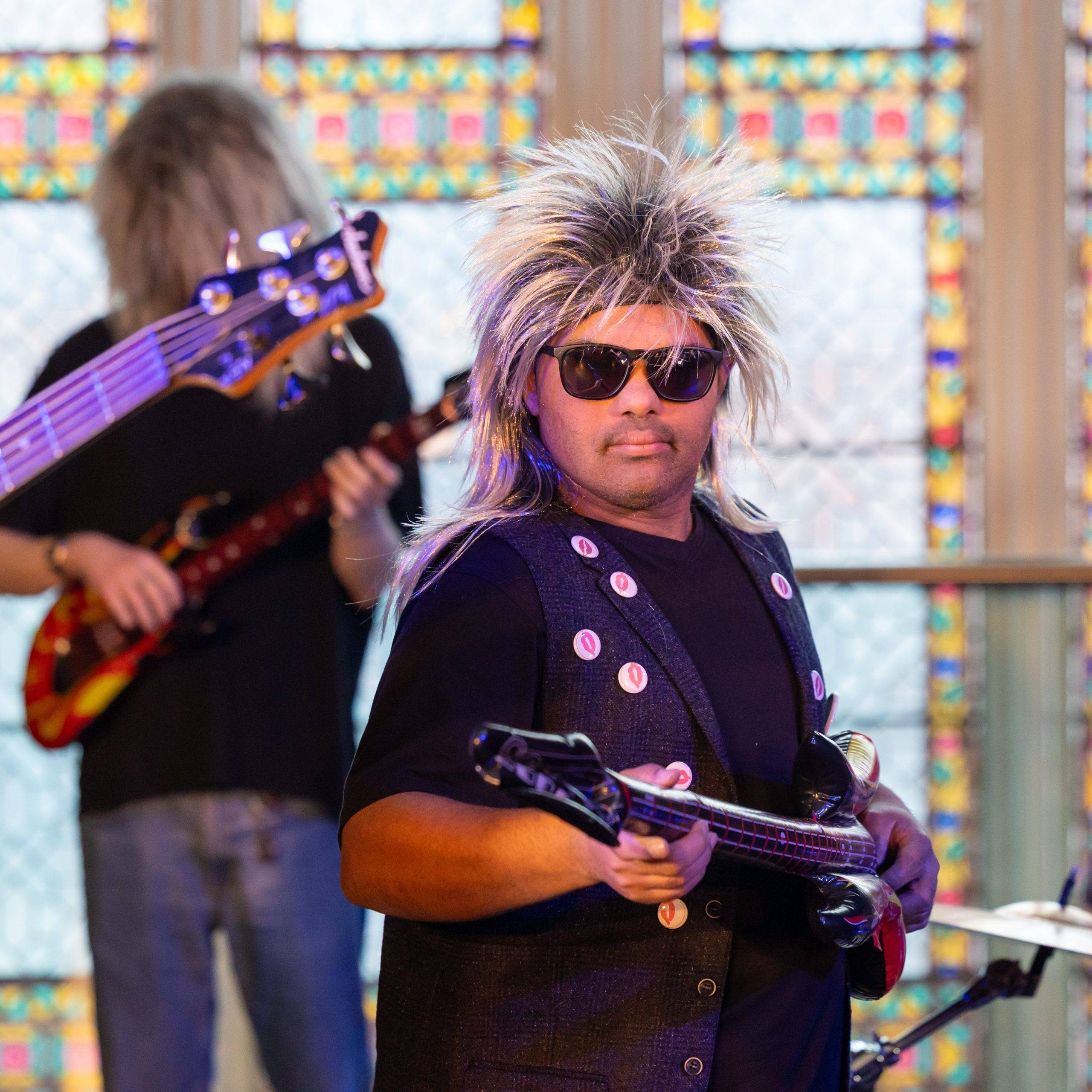 A man stand centre of image wearing a spiky blonde rock wig and sun glasses. He is holding an inflatable guitar. Behind him and out of focus are more band members playing guitars. They are all in front of a bright stained glass window.