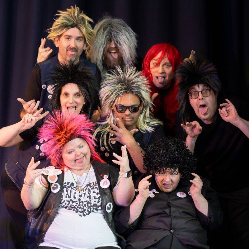 8 people crowd together for a group photo wearing different coloured rock wigs, pulling faces and doing rock hand gestures