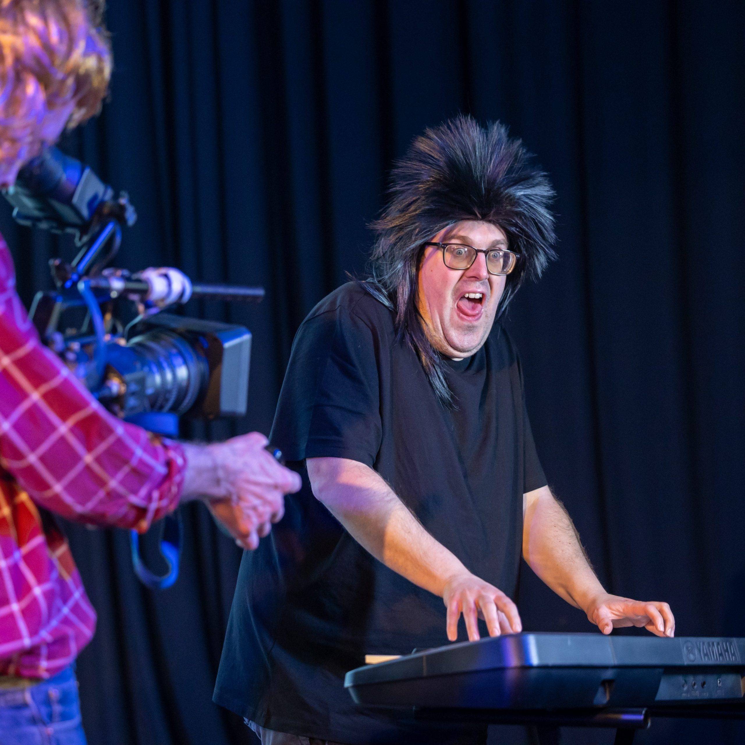 At the centre of the photo is a man wearing a black t-shirt, a black spiky wig and glasses. He is playing a keyboard. In front to the left of the image there is a camera man who is holding a video camera.
